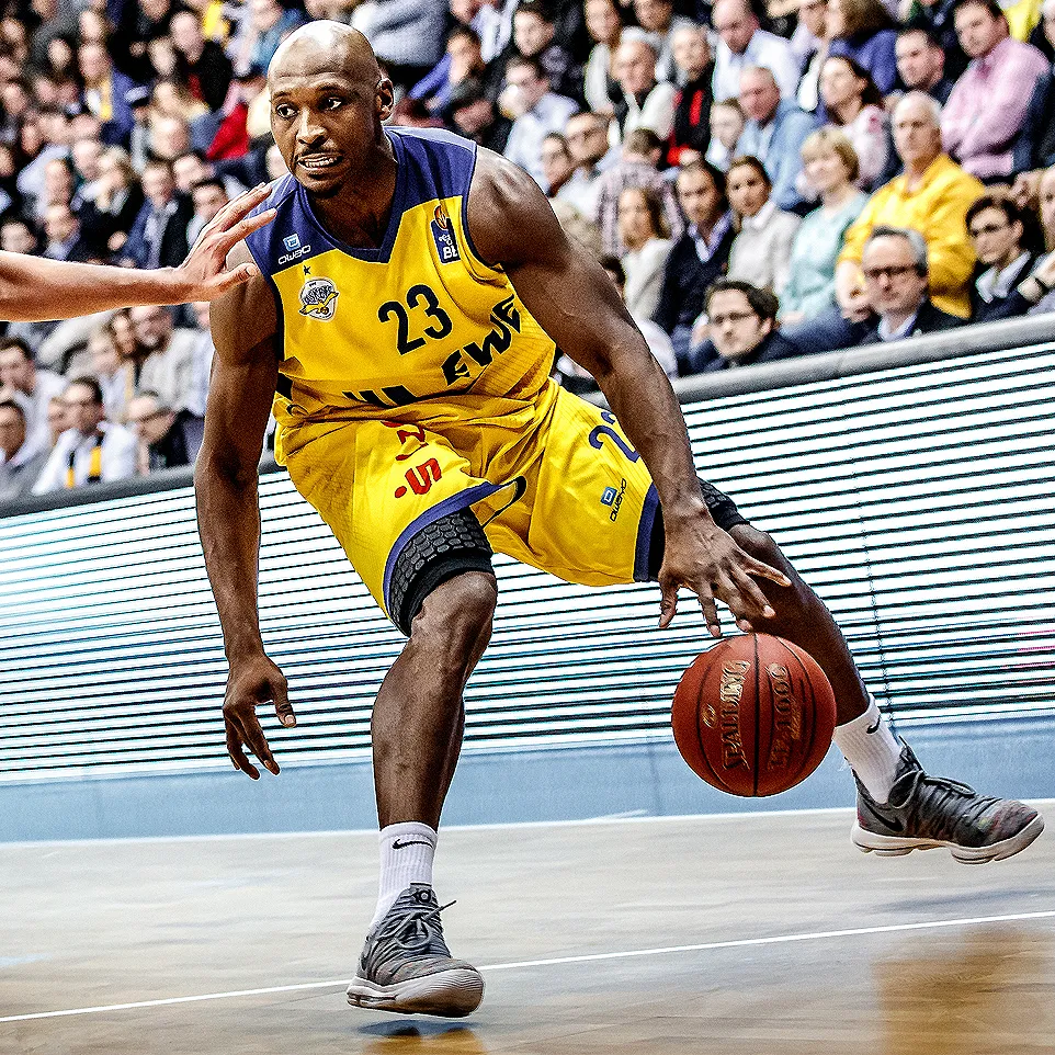 Een basketbalspeler in een wit-blauw owayo-shirt met nummer 4 dribbelt met een bal op een basketbalveld voor een publiek.